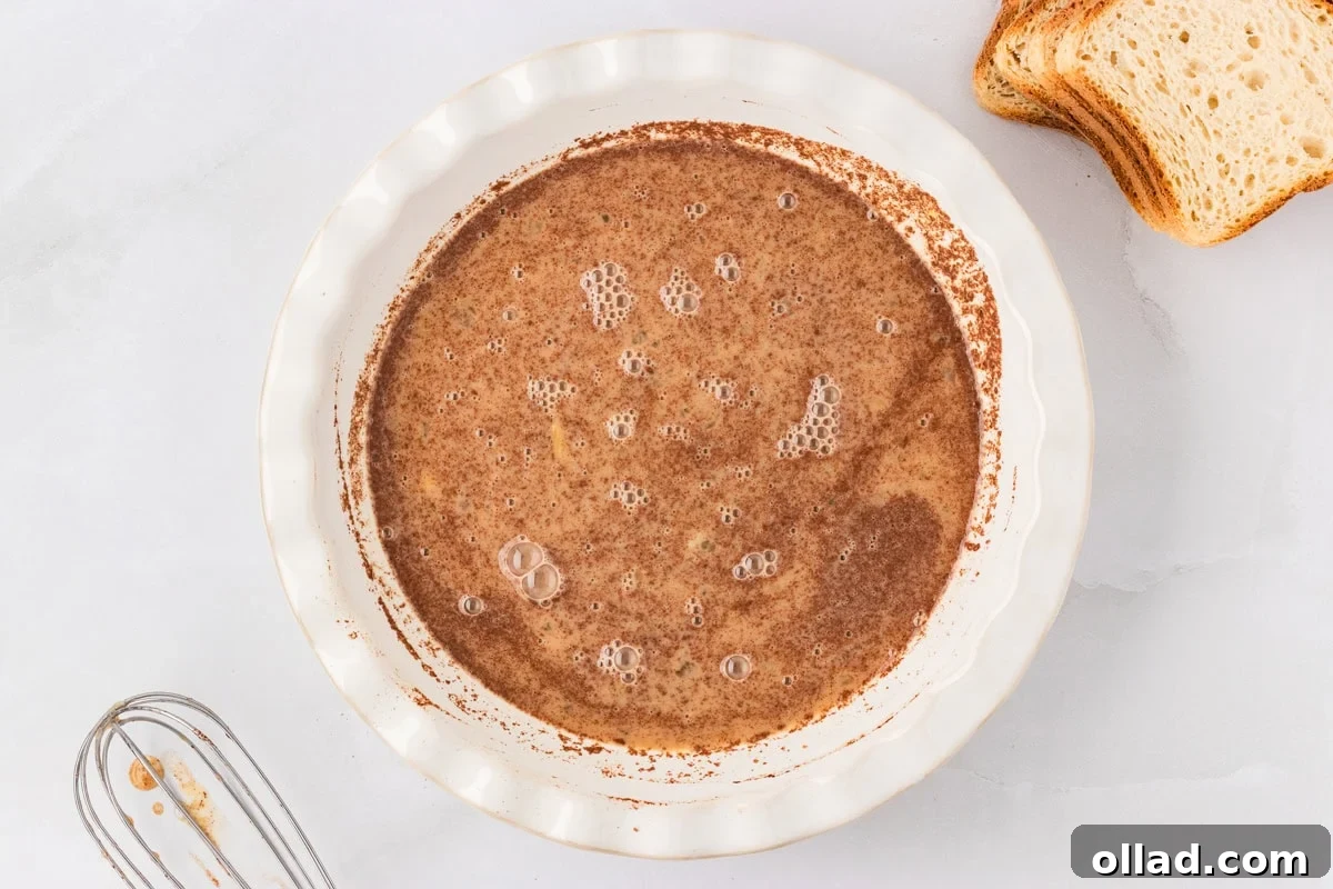An overhead view of a gleaming white shallow pie dish, filled with a creamy, well-whisked egg mixture, visibly speckled with aromatic cinnamon and nutmeg, ready for dipping. Beside it, a neatly stacked pile of gluten-free bread slices awaits its turn to be transformed.