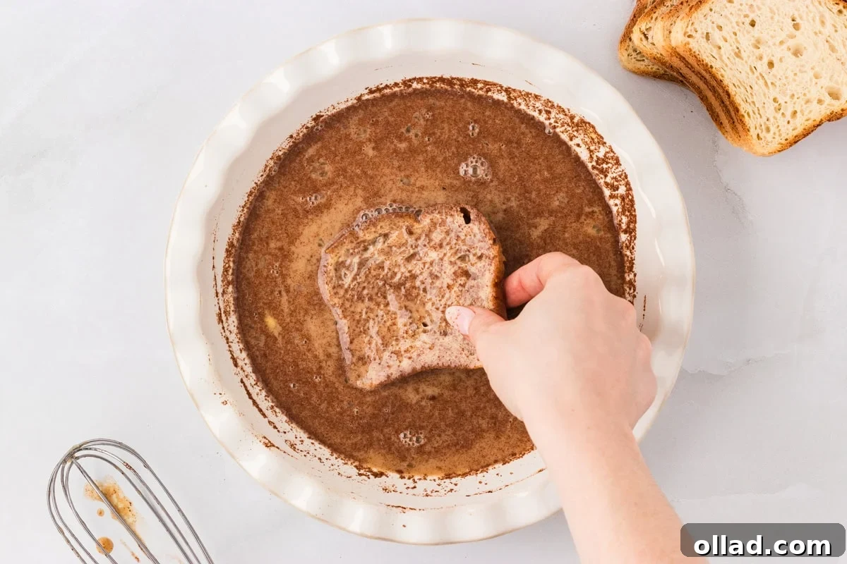 A hand gently lowers a slice of robust gluten-free bread into a shallow dish filled with the creamy, spiced egg mixture. Other slices of bread are stacked nearby, ready to be dipped and transformed into golden French toast.