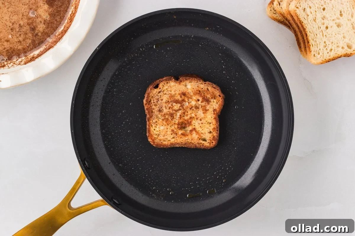 A single slice of Gluten-Free French Toast sizzling gently in a nonstick skillet, achieving a perfect golden-brown hue on one side as it cooks evenly.