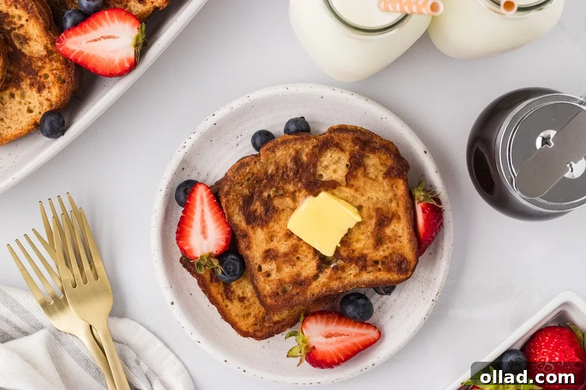 A pristine white plate holds a stack of freshly cooked Gluten-Free French Toast, glistening with a pat of butter, and surrounded by a vibrant medley of fresh strawberries and blueberries. In the soft-focus background, a glass of milk and a bottle of maple syrup complete the inviting breakfast scene.