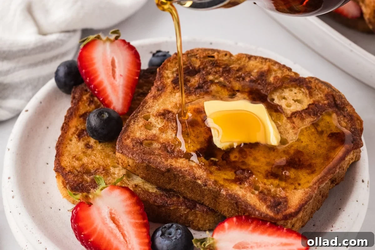 A close-up shot of a single slice of warm Gluten-Free French Toast on a white plate, topped with a melting pat of butter and drenched in a thick stream of golden maple syrup. Scattered fresh berries provide a burst of color and freshness.