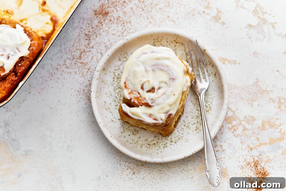 Irresistible Gluten-Free Cinnamon Rolls 22 A frosted gluten-free cinnamon roll on a speckled ceramic plate with a fork beside it.