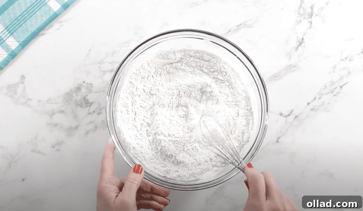 Overhead view of a person whisking a mixture of dry ingredients—gluten-free flour, sugar, and yeast—in a large glass mixing bowl on a white marble surface.