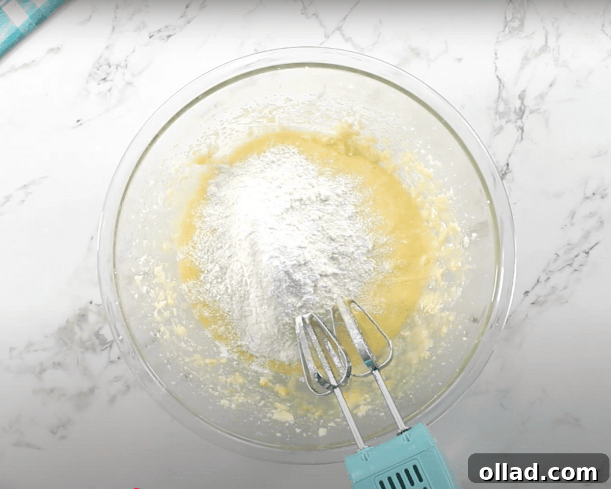 The dry ingredient mixture is being added to the wet ingredients in the glass mixing bowl, with a blue hand mixer resting nearby.