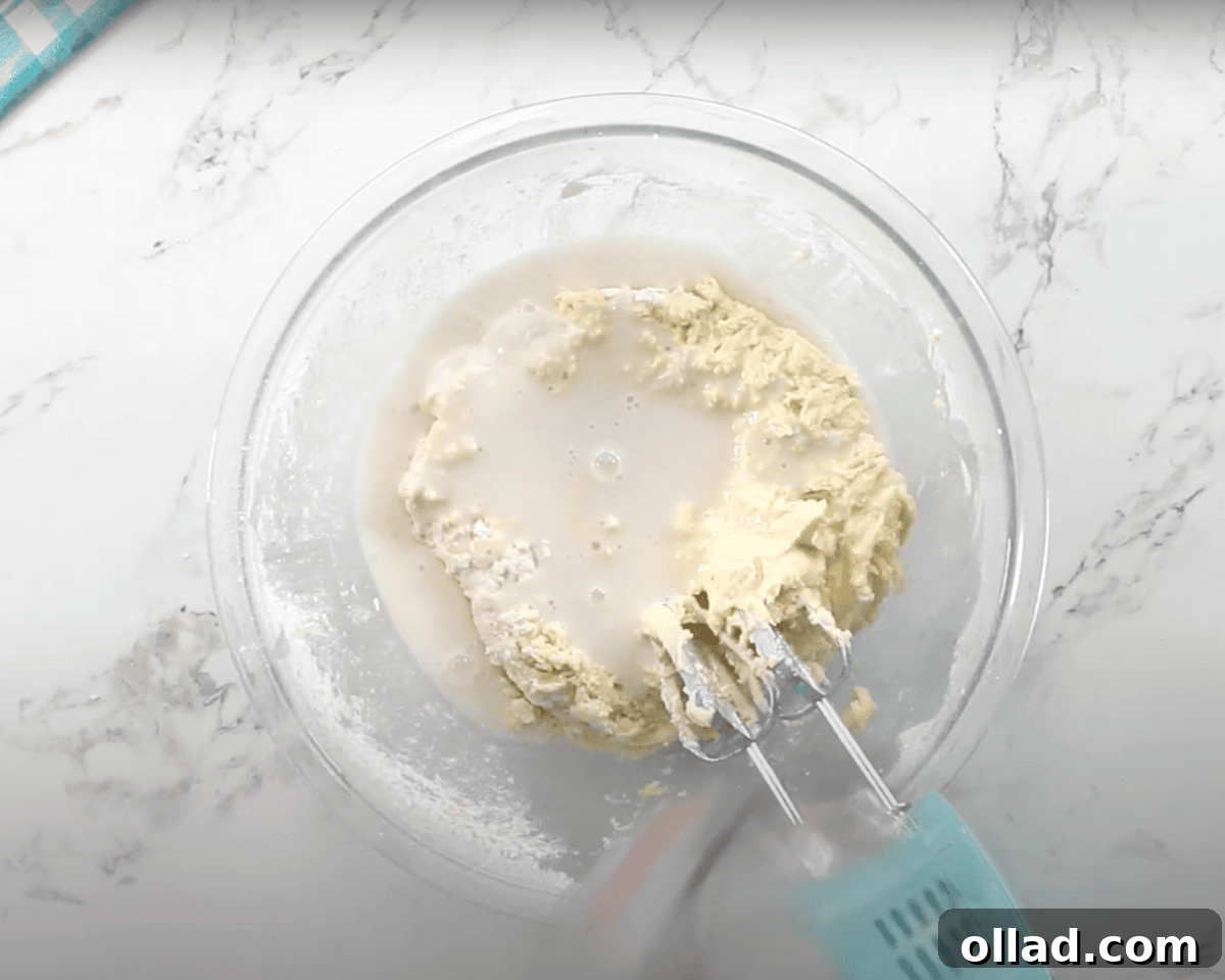 The batter is partially mixed, and a liquid, likely milk or buttermilk, is being added to the bowl.