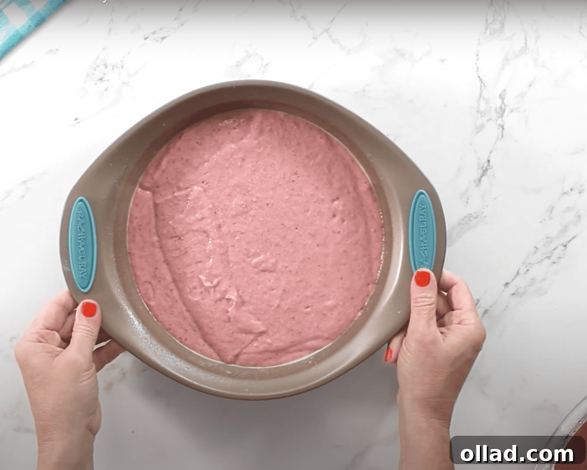 A round cake pan filled with smooth, pink strawberry cake batter. A person with red nail polish holds the pan steady on a white marble surface.