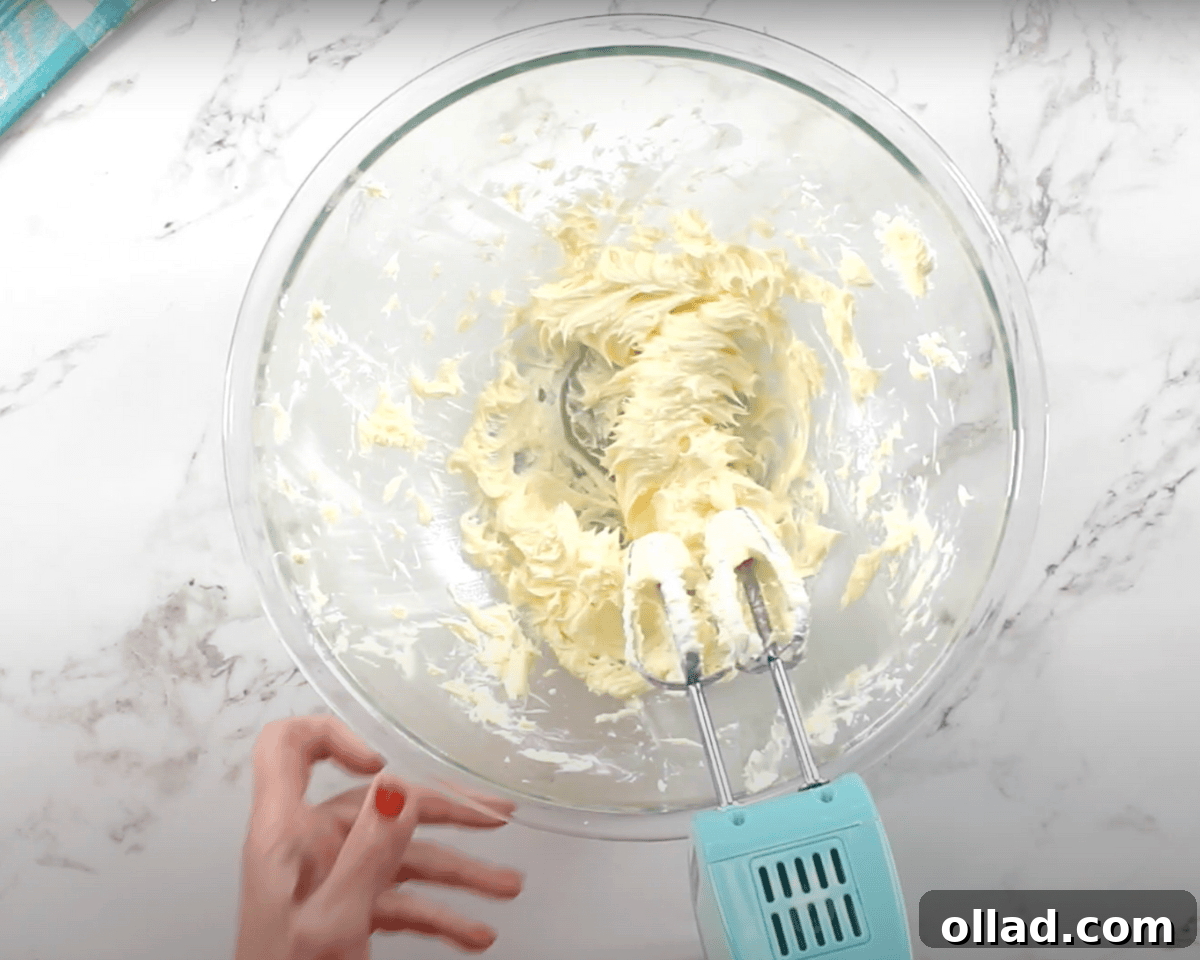 A glass mixing bowl with softened butter being whipped with a blue hand mixer, creating a smooth and fluffy consistency.