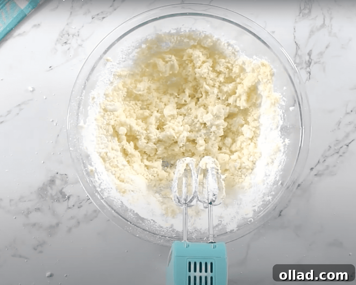A glass mixing bowl with powdered sugar added to the whipped butter, forming a crumbly mixture before being fully blended.