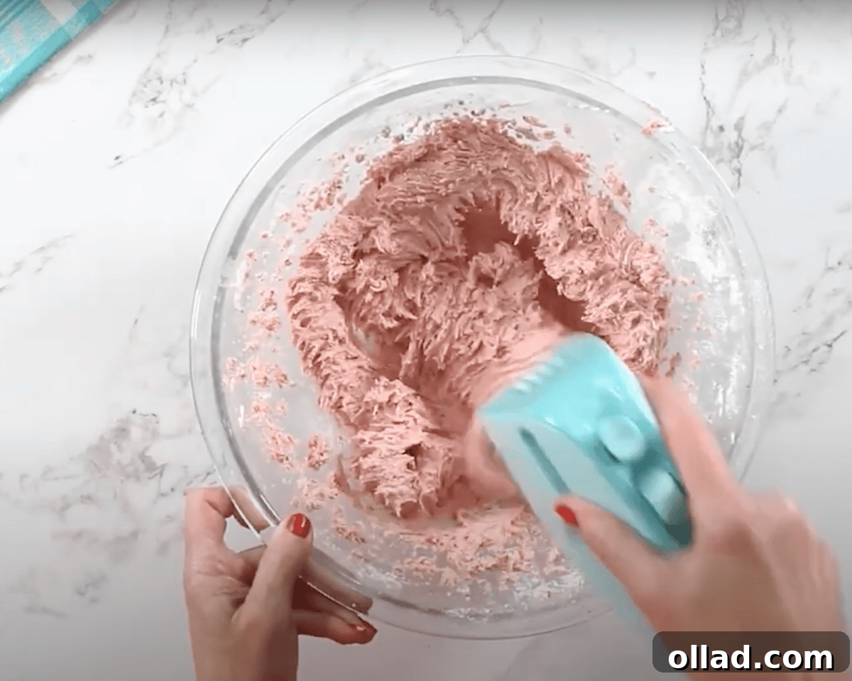 A glass mixing bowl with a pink strawberry buttercream frosting being mixed with a blue hand mixer. The frosting has a fluffy and creamy texture.