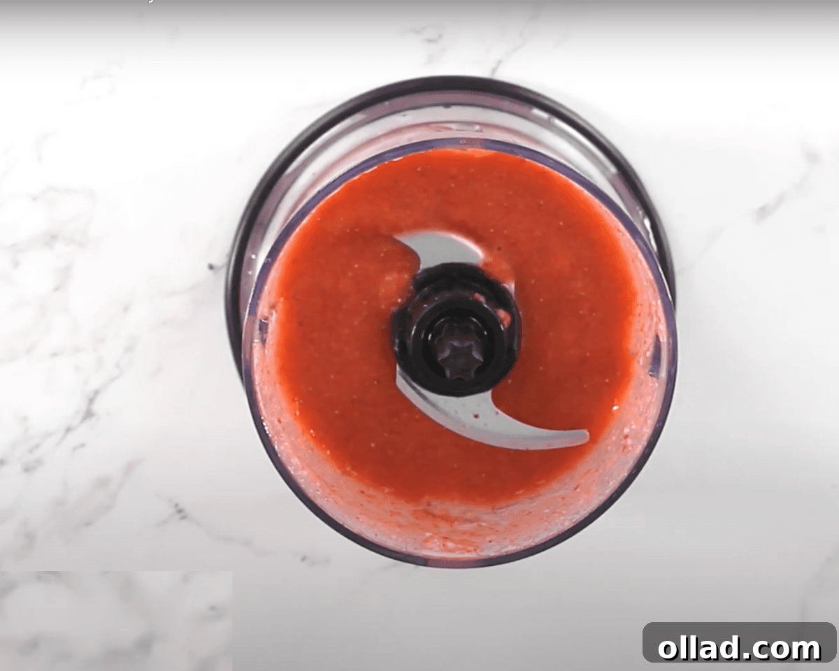 Overhead view of a food processor filled with smooth strawberry puree, sitting on a white marble countertop.