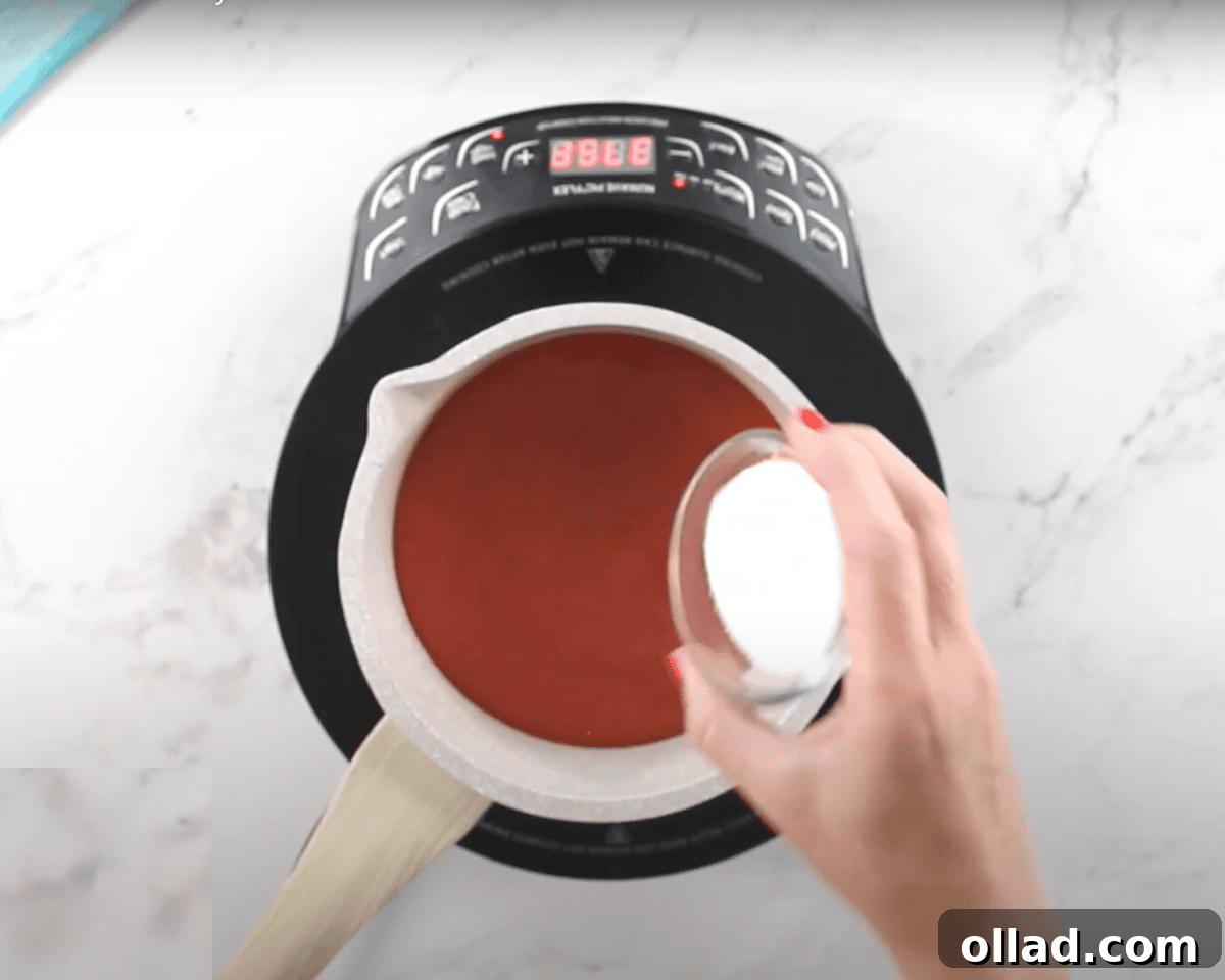 A saucepan filled with strawberry puree on an induction cooktop. A hand is adding granulated sugar to the mixture.
