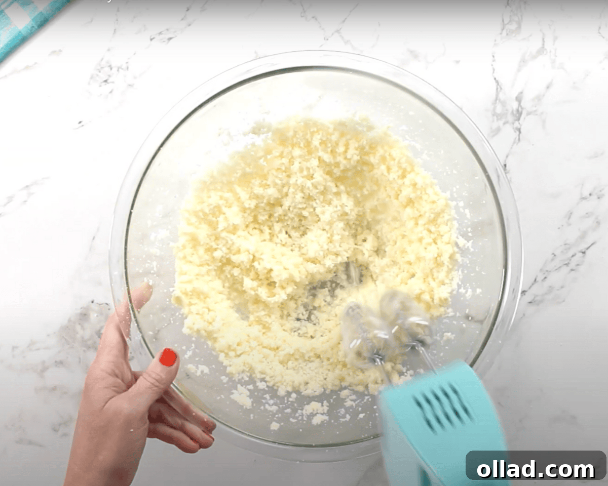 A glass mixing bowl with butter and sugar being mixed with a hand mixer. The mixture appears crumbly and pale yellow.