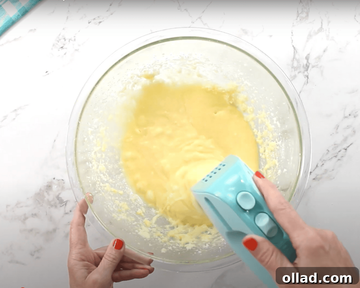 A glass mixing bowl with a smoother, yellow cake batter being mixed with a blue hand mixer.
