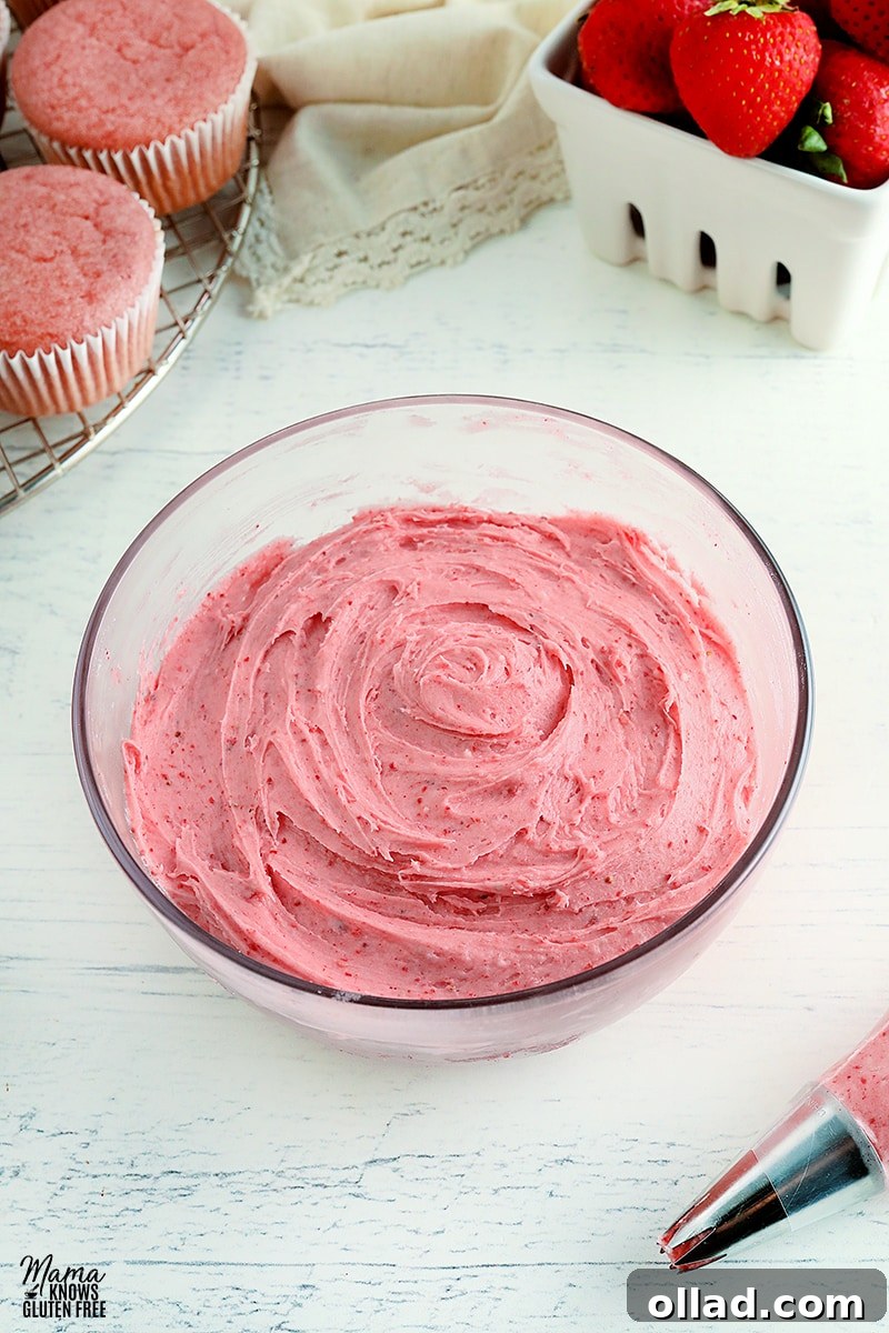 A glass bowl filled with luscious strawberry frosting, surrounded by fresh strawberries and decorated cupcakes