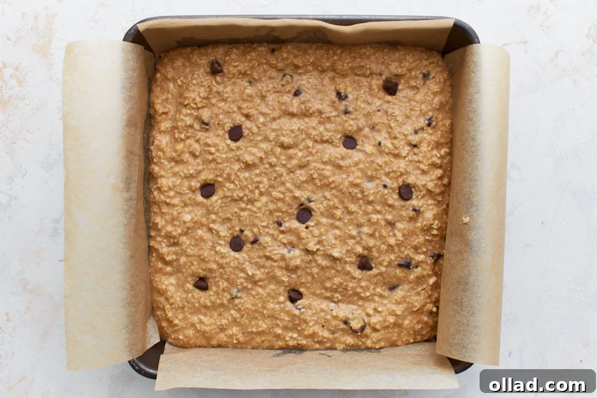 Chocolate chip oatmeal batter spread evenly in a parchment-lined square baking pan before baking.