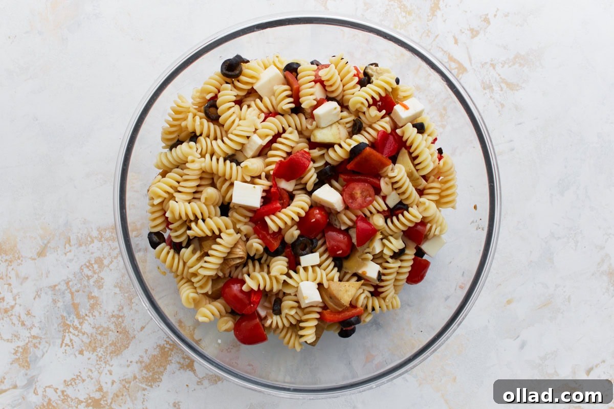 A clear glass mixing bowl filled with a colorful array of prepared ingredients: cooked rotini pasta, halved cherry tomatoes, sliced black olives, vibrant roasted red peppers, chopped artichoke hearts, and cubed mozzarella cheese, awaiting the dressing.