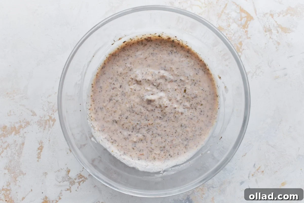 A small, clear glass bowl holding a freshly prepared homemade creamy Italian dressing, flecked with green herbs, perfectly smooth and ready to be poured over a pasta salad.