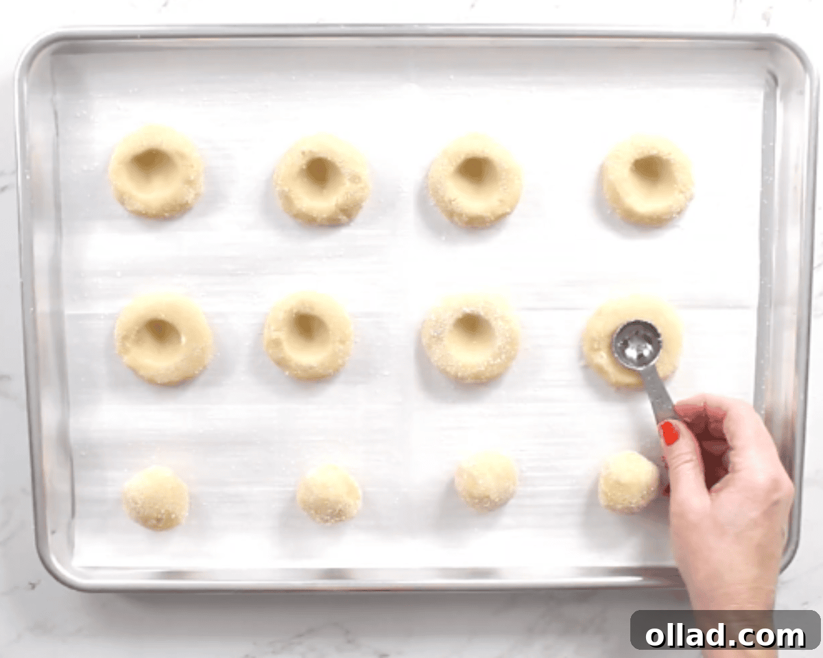 A gluten-free thumbprint cookie on a cookie sheet with a teaspoon being used to create a small indent for the jam.
