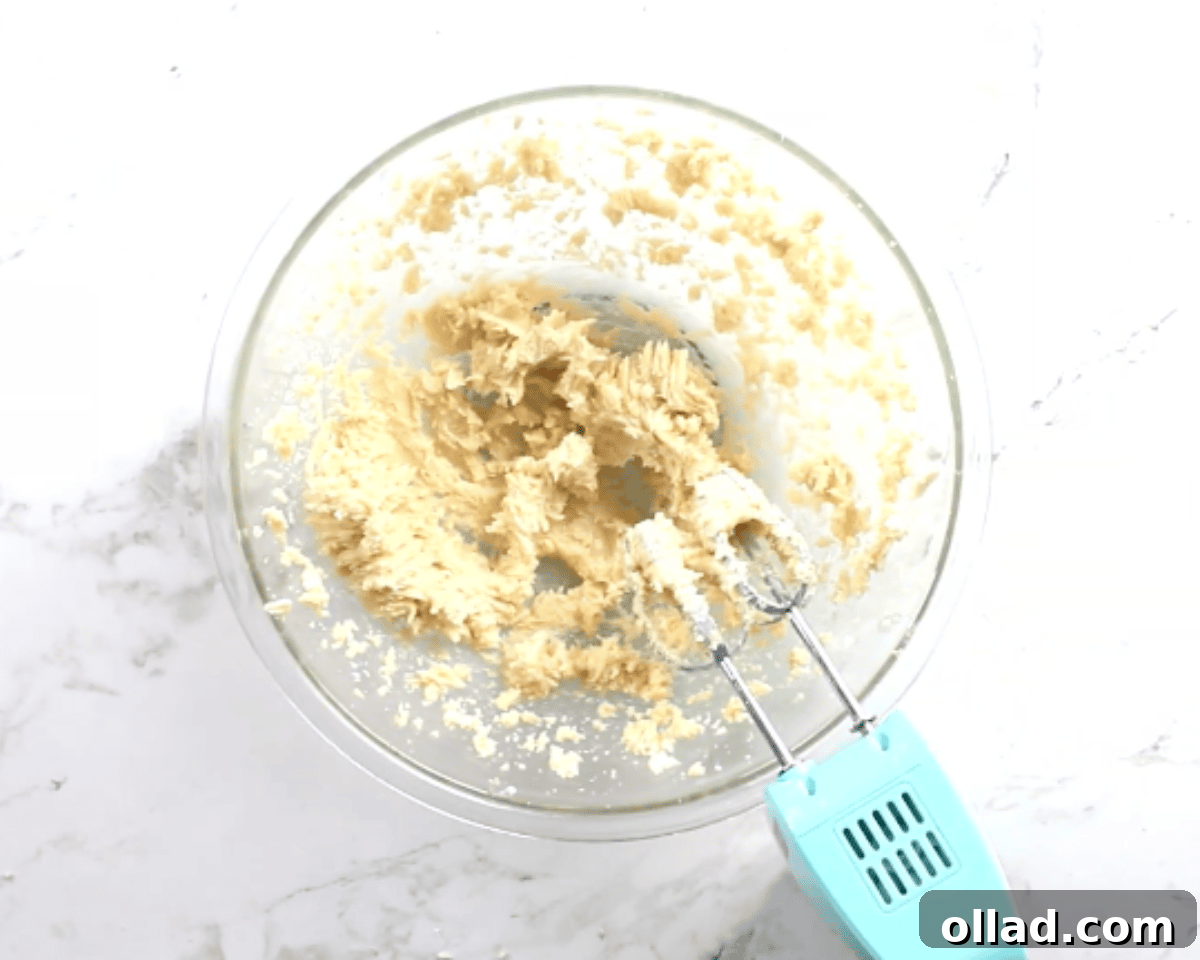 Creamed butter and sugars in a glass mixing bowl with an electric hand mixer on a marble countertop.