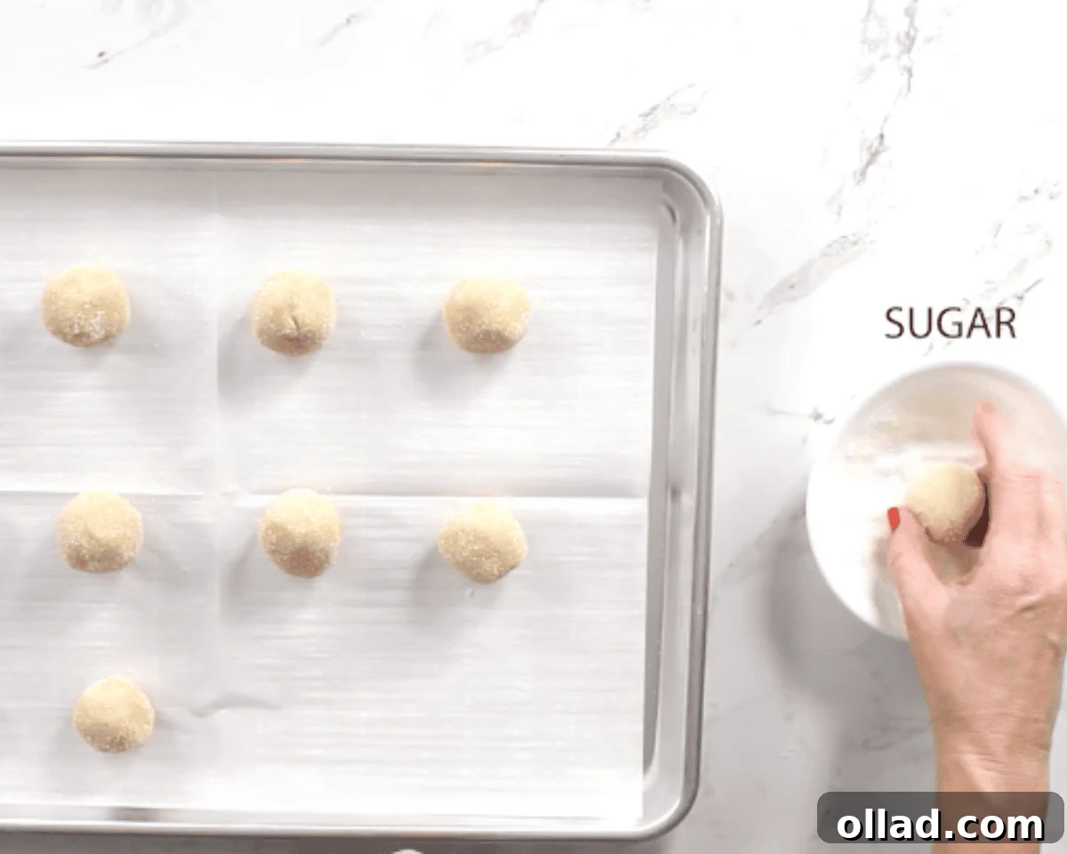Gluten-free thumbprint cookie dough balls being rolled in granulated sugar on a cookie sheet.