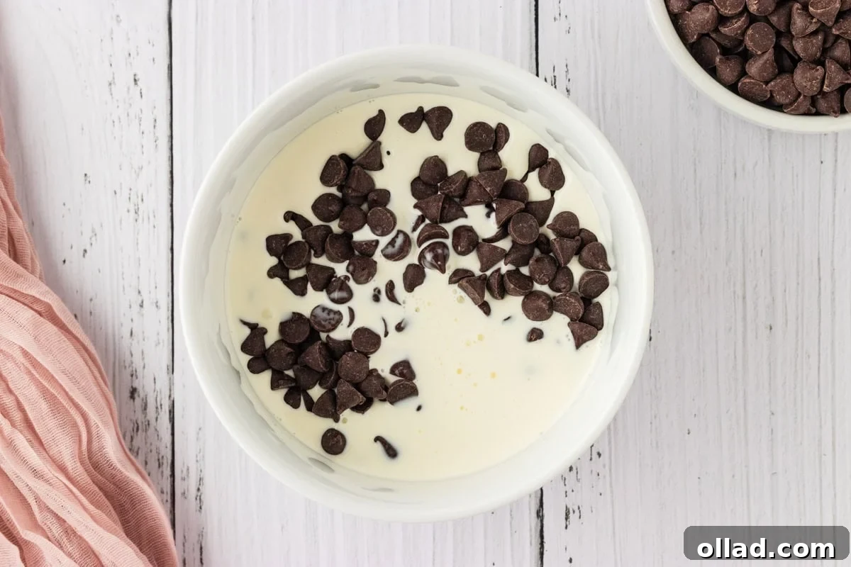 A medium white bowl filled with creamy heavy cream and semi-sweet chocolate chips, prepared for melting to create the ganache.
