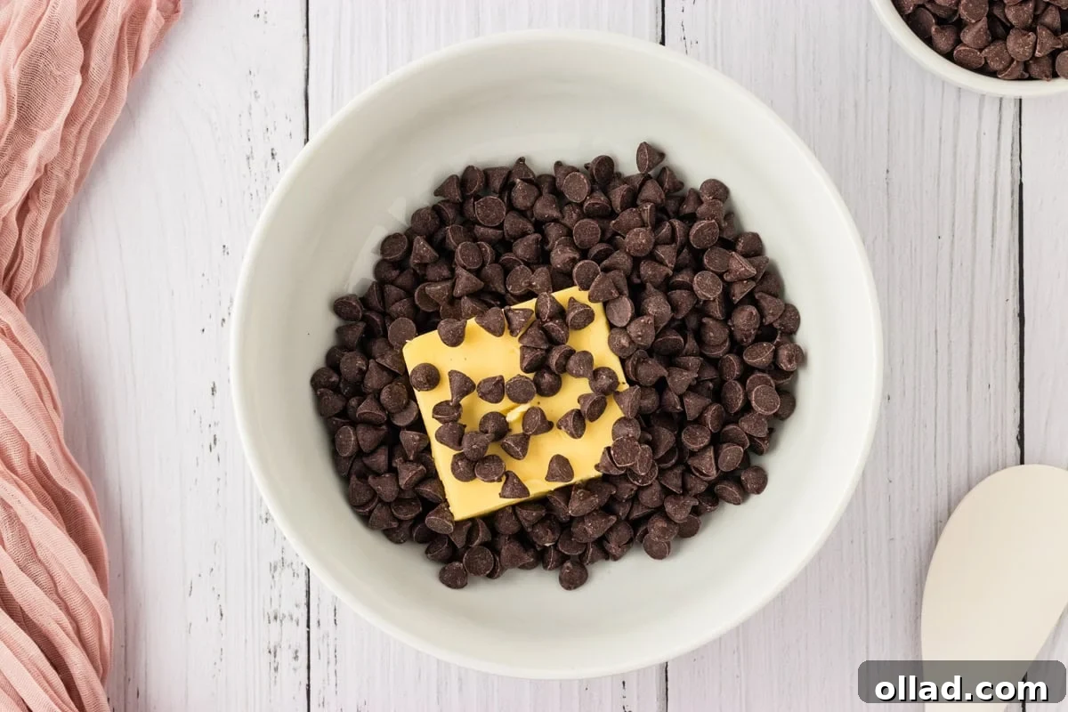 A white ceramic bowl containing rich semi-sweet chocolate chips and a block of unsalted butter, ready to be melted together.