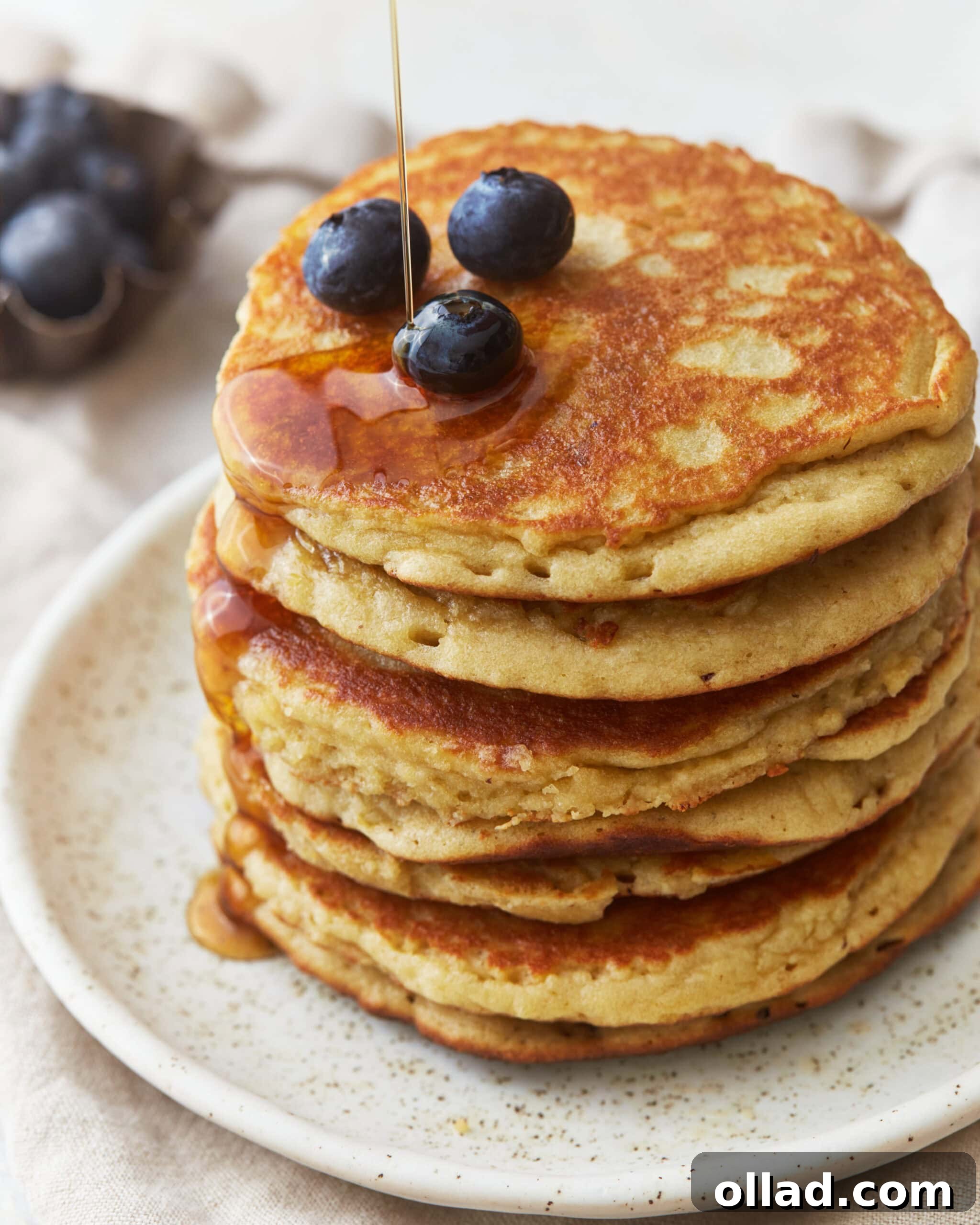 A beautifully arranged stack of golden almond flour pancakes on a plate, adorned with fresh blueberries and glistening maple syrup, photographed from a three-quarters perspective.