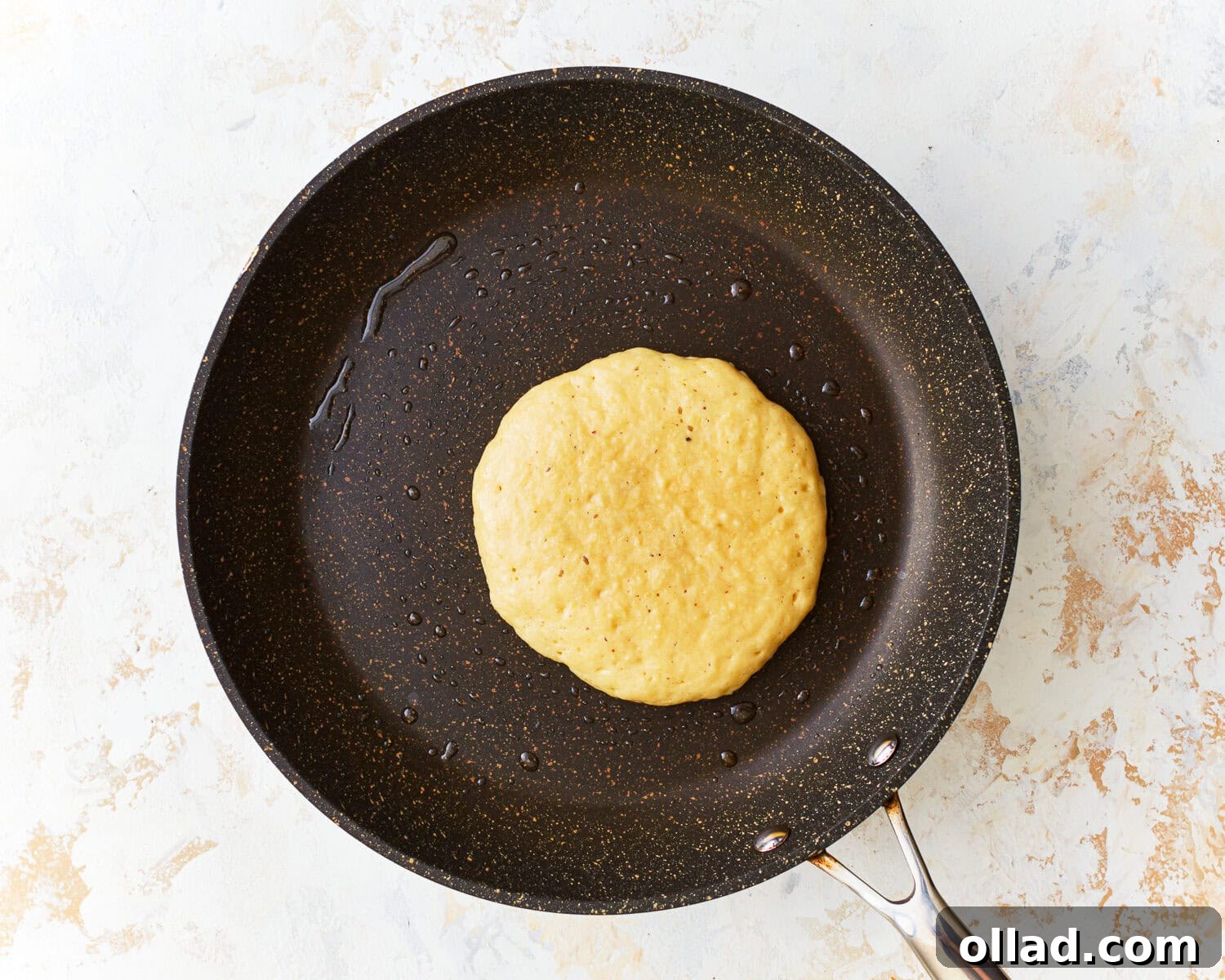 Almond flour pancake batter being carefully poured from a scoop into a hot, oiled frying pan, forming round pancakes.