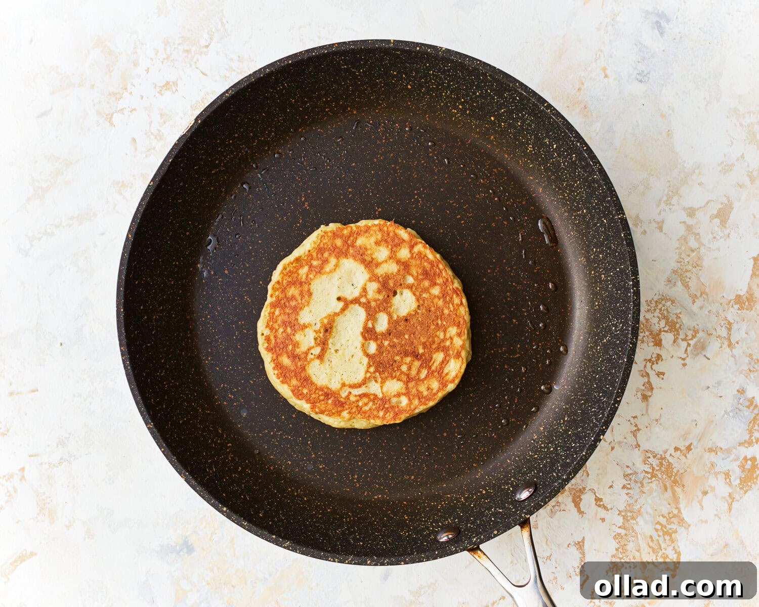 A single almond flour pancake sizzling in a frying pan, golden brown on one side and lightly bubbling on the uncooked side, ready to be flipped.