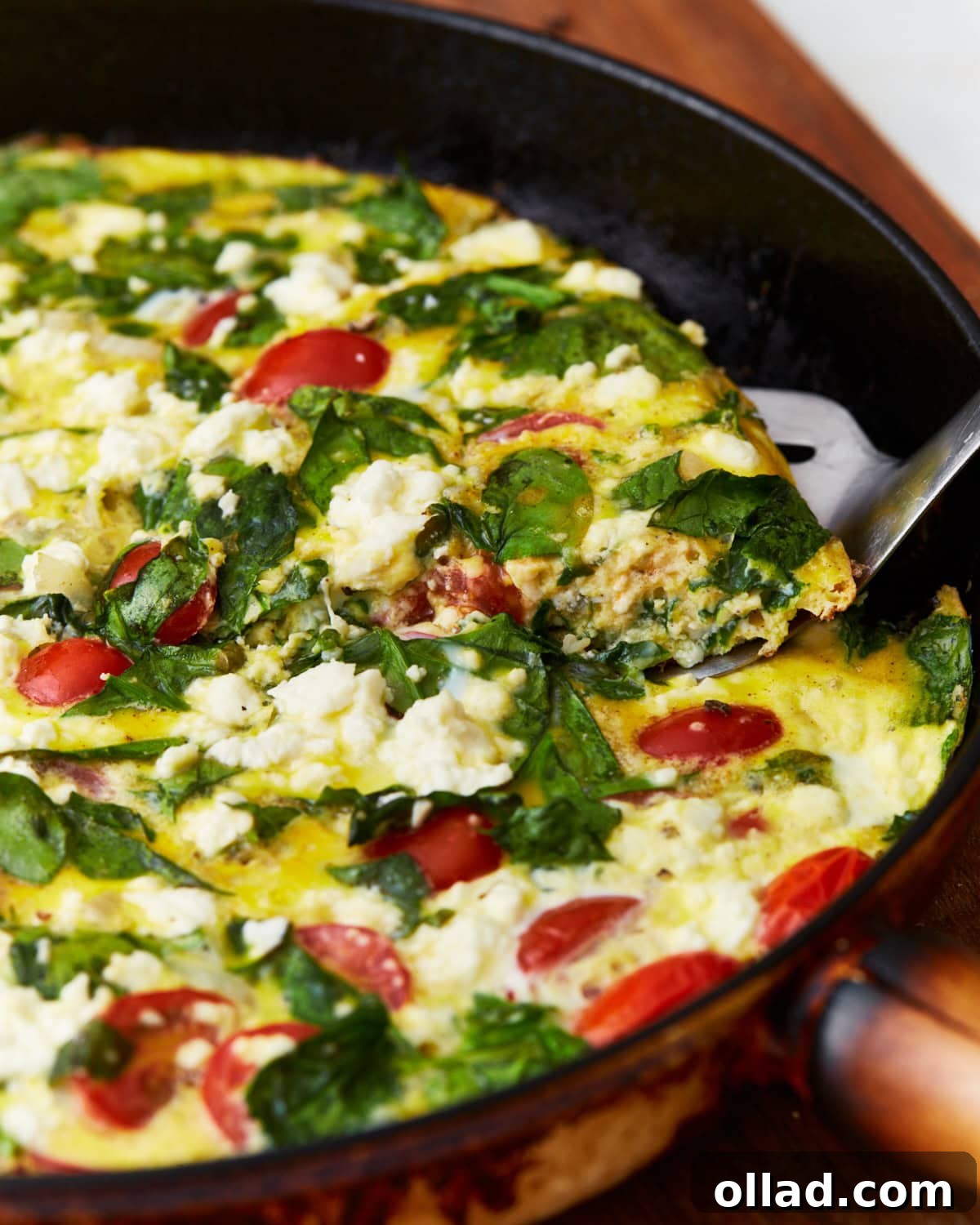 A vibrant slice of frittata, rich with tomatoes, spinach, and feta cheese, being expertly lifted from a black skillet with a metal spatula.