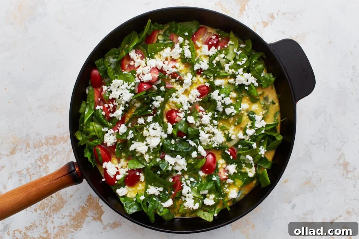 A cast iron skillet holds a colorful, uncooked frittata mixture, generously topped with fresh spinach, bright cherry tomatoes, and crumbled feta cheese, awaiting its transformation.