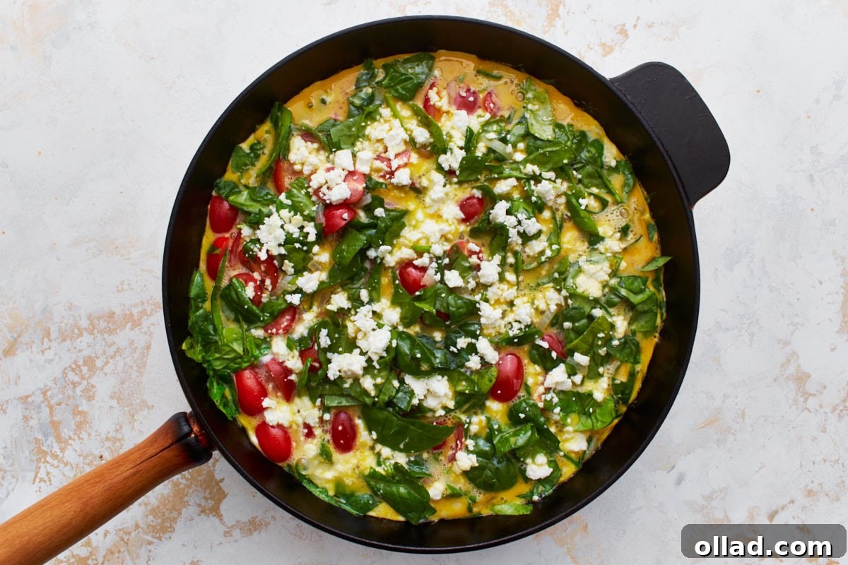 A cast iron skillet displays a frittata that has begun to set around the edges, showcasing the vibrant spinach, cherry tomatoes, and crumbled feta cheese, ready for the oven.