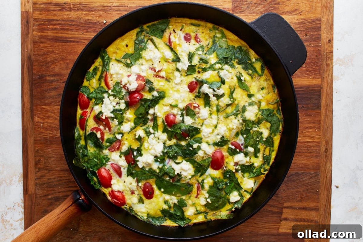 A beautifully baked frittata, brimming with spinach, tomatoes, onions, and crumbled feta cheese, rests in a black cast-iron skillet, fresh from the oven and perfectly golden.