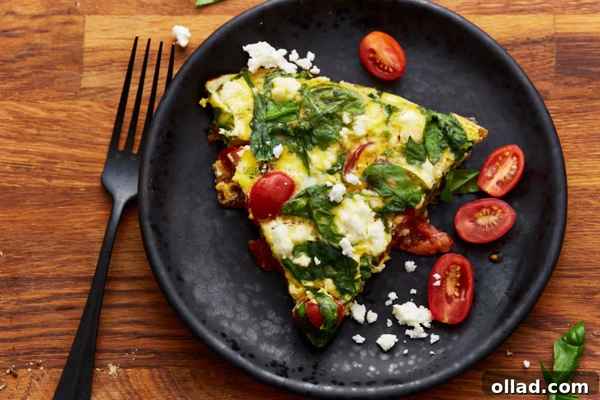 A beautifully plated slice of baked frittata, adorned with fresh spinach, bright tomatoes, and creamy feta cheese, served on a sleek black plate with a matching black fork, set against a warm wooden backdrop.