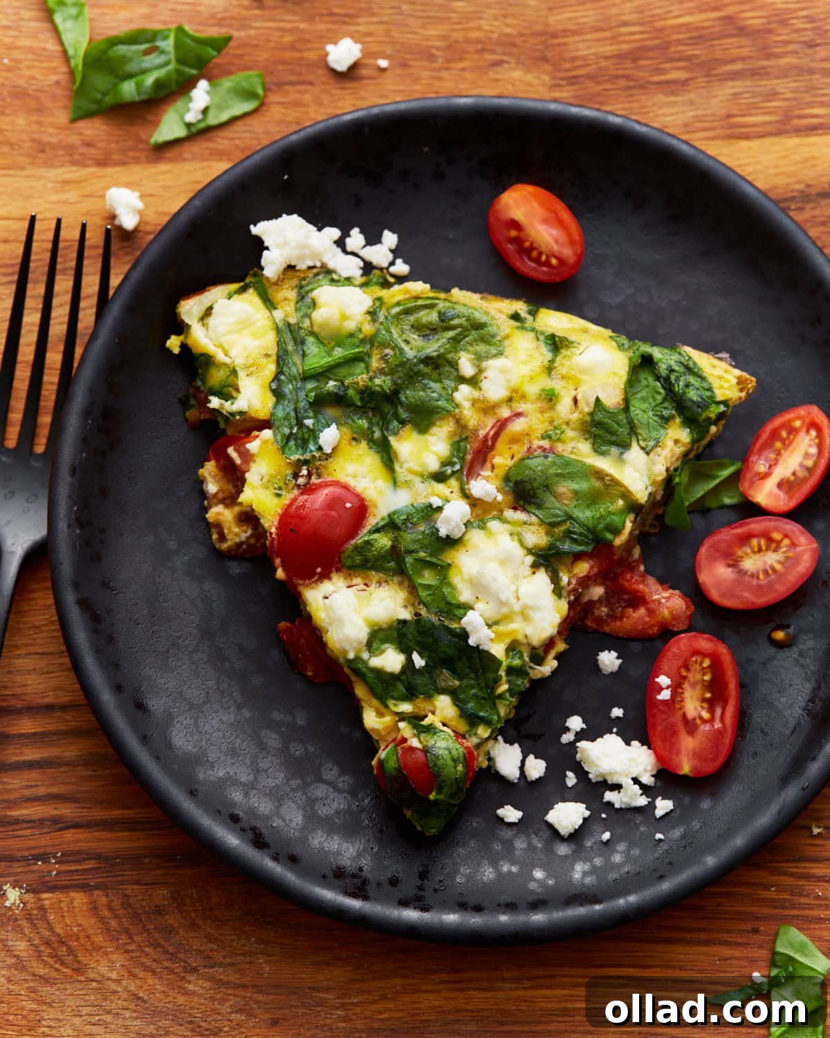 A delectable slice of baked frittata, beautifully topped with fresh spinach, juicy tomatoes, and creamy feta cheese, rests on a black plate with a matching black spoon, all situated on a rustic wooden surface.