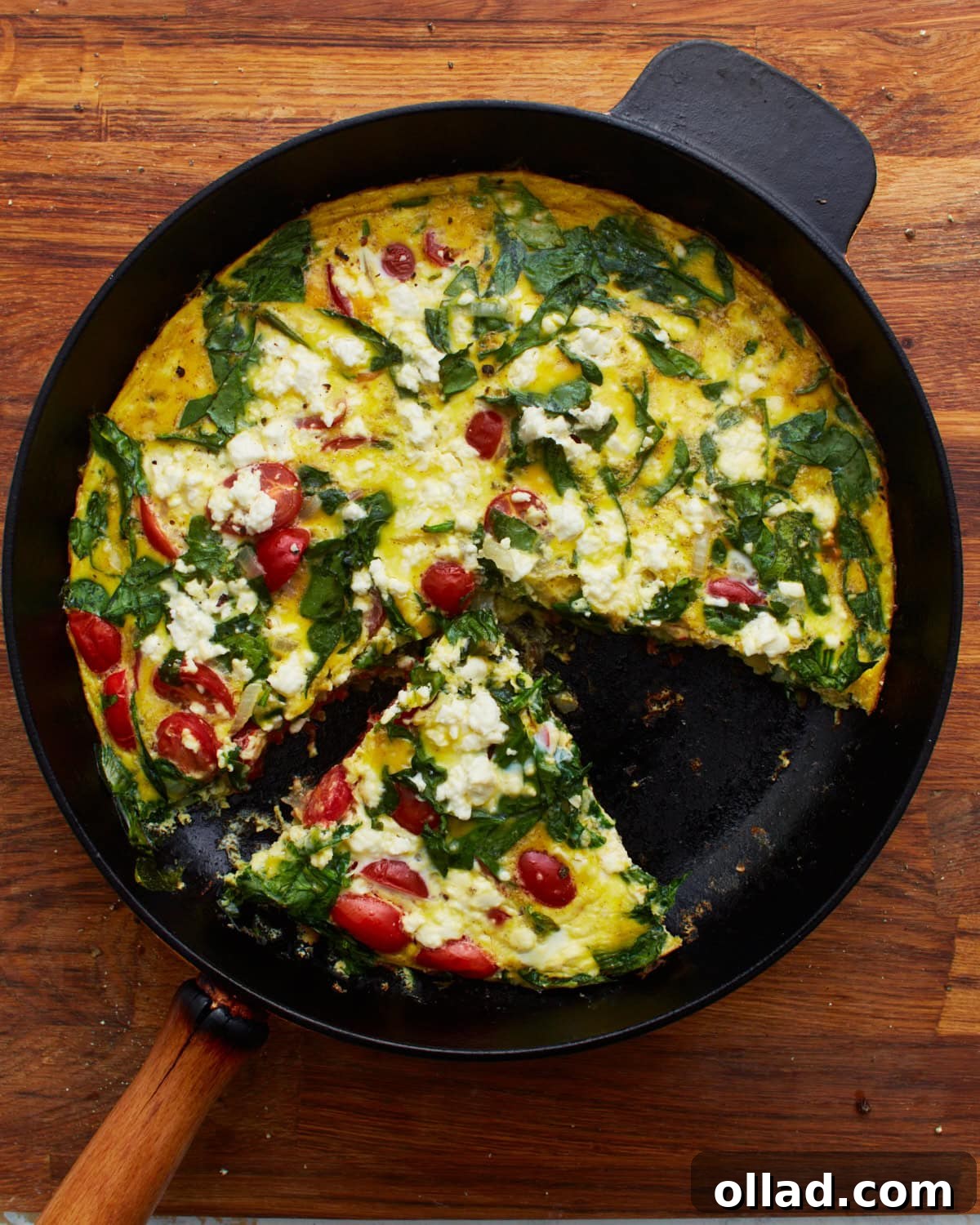 A freshly baked frittata, topped with vibrant spinach, halved grape tomatoes, and crumbled feta cheese, presented in a black cast-iron skillet. A single slice has been removed, highlighting the delicious layers, with the skillet resting on a rustic wooden surface.