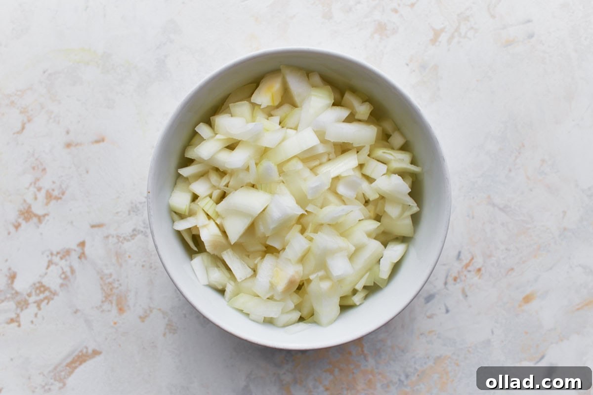 A small white bowl filled with precisely diced yellow onion, ready for cooking, sits on a clean, light-colored surface.