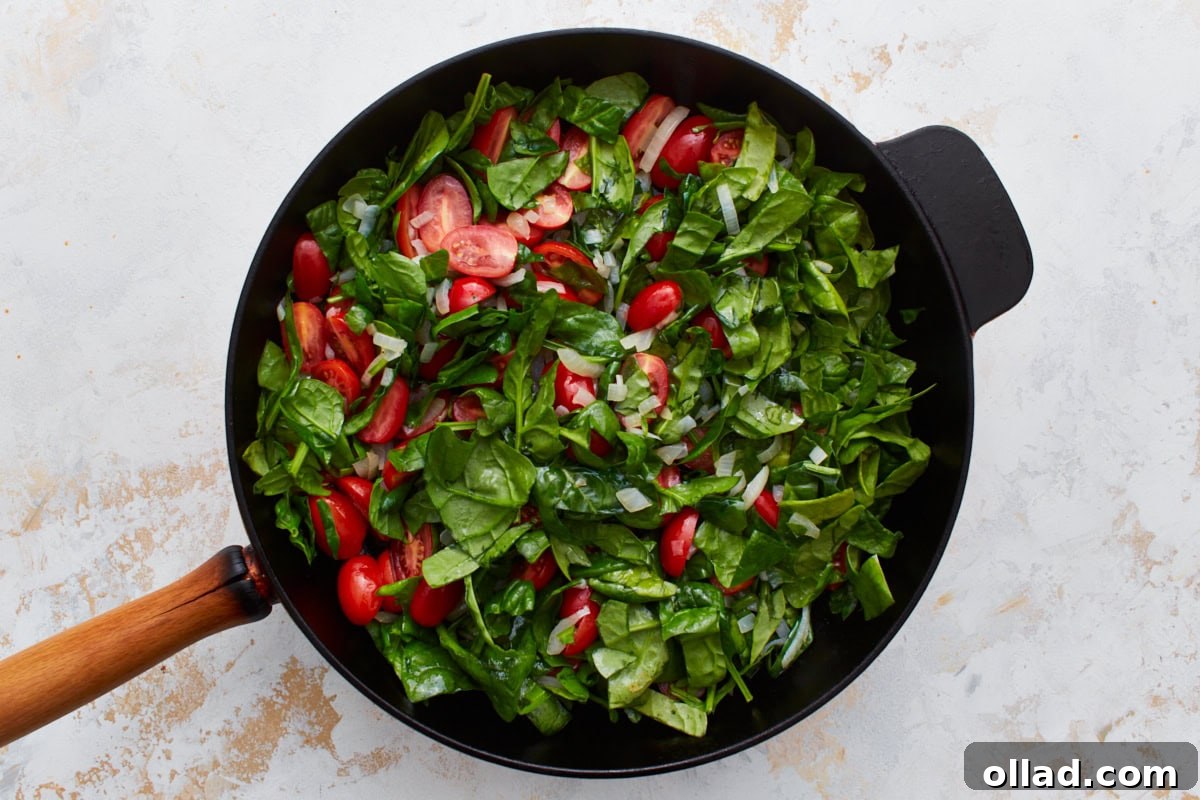 Freshly chopped spinach and vibrant halved grape tomatoes have been added to a black cast-iron skillet, creating a colorful and healthy mix.