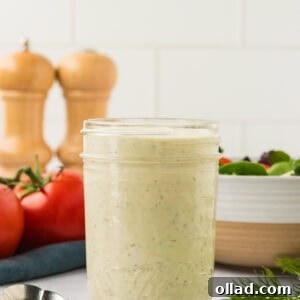A glass mason jar filled with creamy dairy-free ranch dressing, set on a white countertop with tomatoes, fresh dill, and a spoon nearby.
