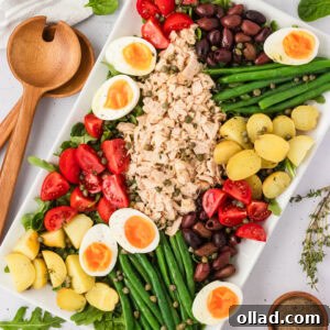 Overhead view of a beautifully arranged Tuna Nicoise Salad on a white rectangular platter with eggs, tomatoes, olives, green beans, potatoes, and tuna.