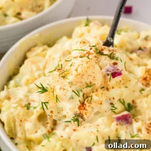 A close-up of a spoonful of creamy gluten-free potato salad with diced potatoes, red onion, and fresh dill, sprinkled with paprika.