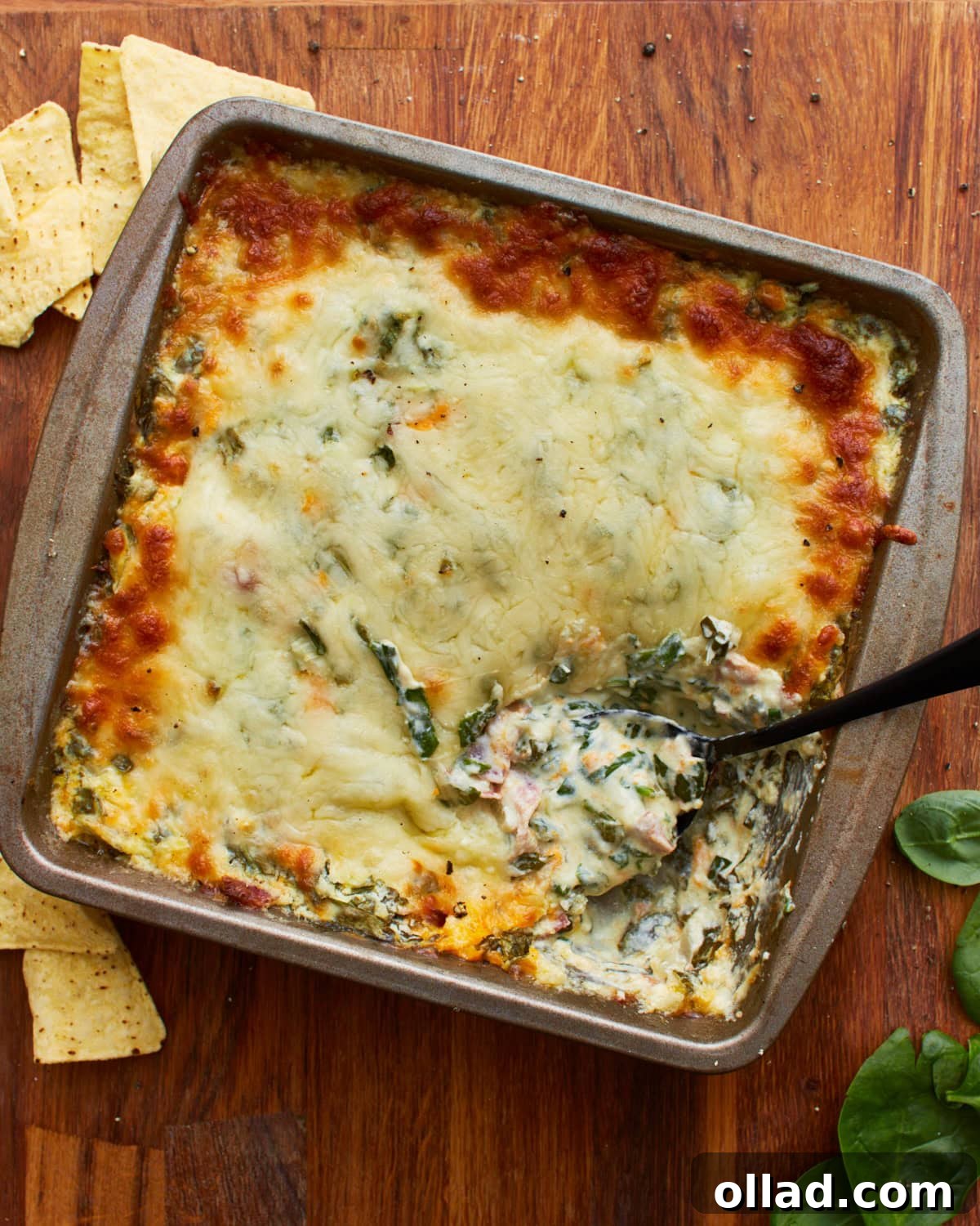 Overhead shot of a spoon dipped into a tin square baking dish with hot spinach dip topped with mozzarella cheese on a wooden surface