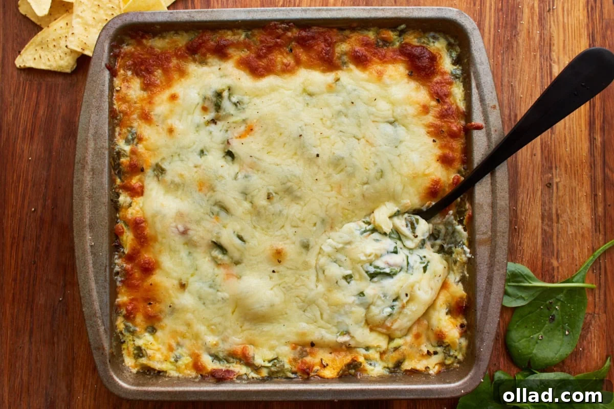 Baked hot spinach dip in a square tin baking dish topped with shredded mozzarella cheese with a spoon dipping in