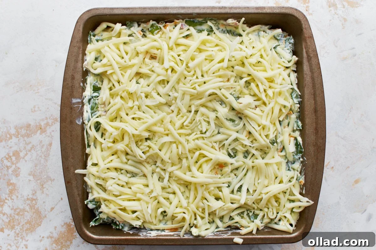 Uncooked hot spinach dip in a square tin baking dish topped with shredded mozzarella cheese