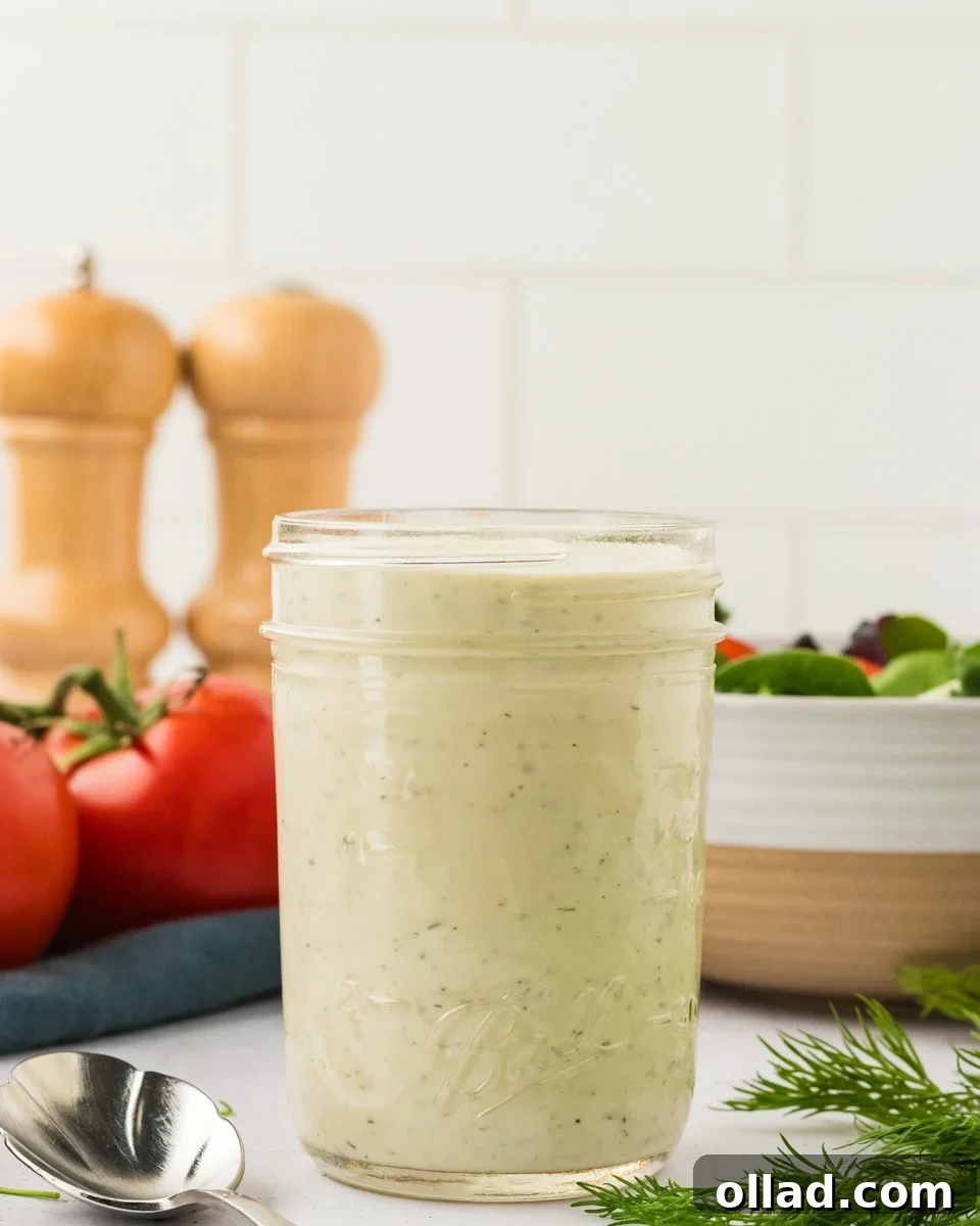 A glass mason jar filled with creamy dairy-free ranch dressing, set on a white countertop with tomatoes, fresh dill, and a spoon nearby.