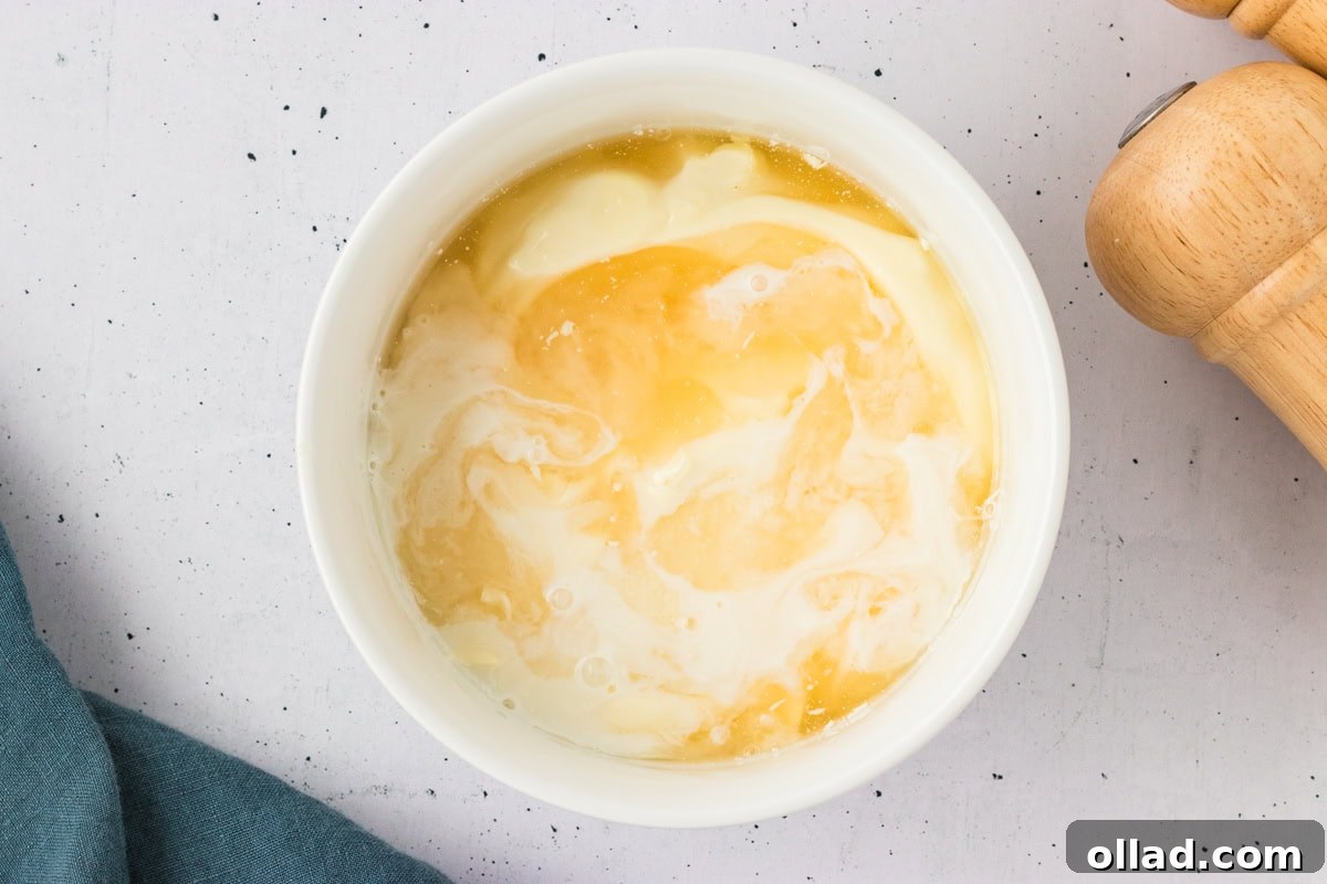 Overhead view of a white bowl with mayonnaise and vinegar beginning to mix together on a speckled white surface.