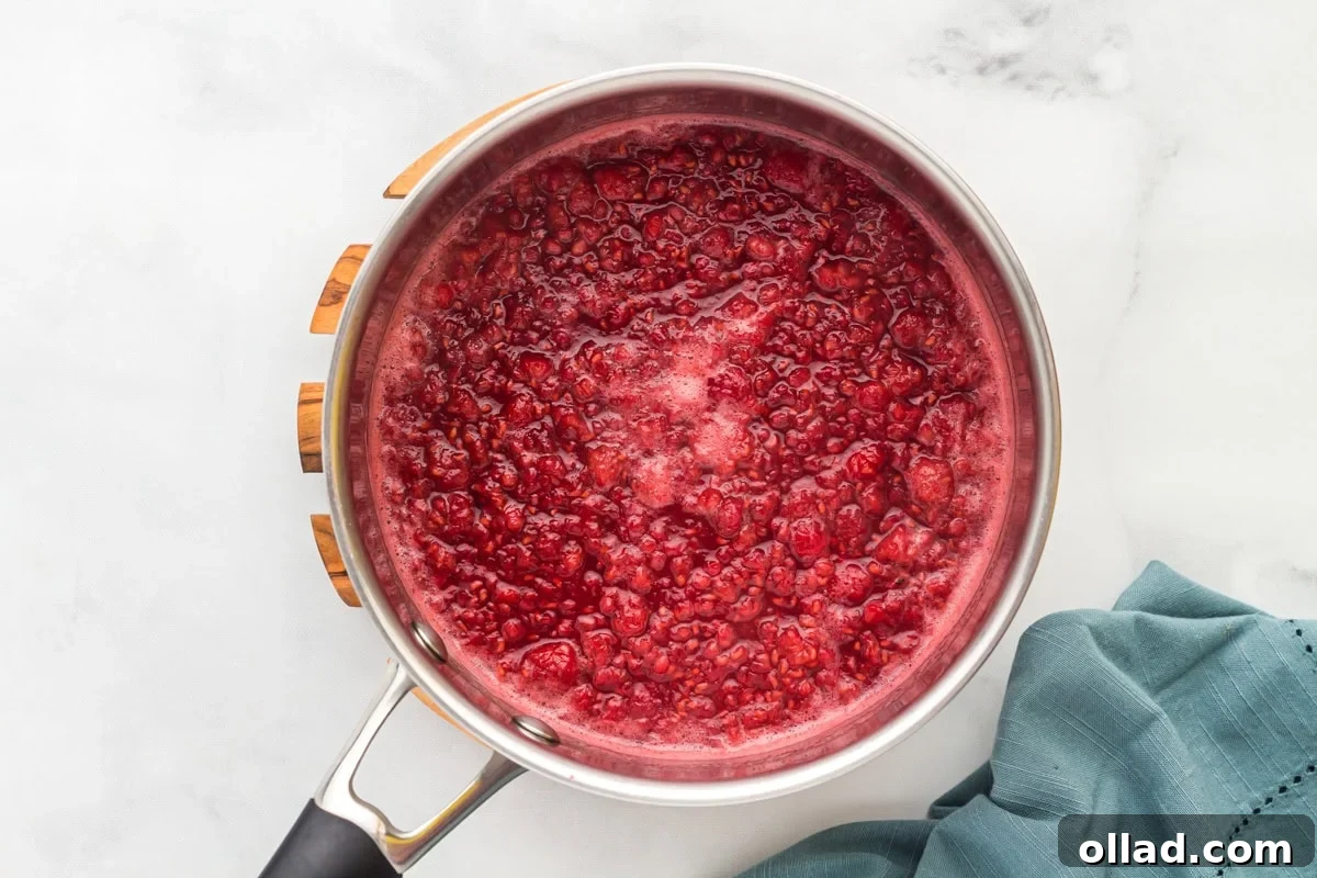 Overhead view of a saucepan filled with simmering raspberries, sugar, lemon juice, and water, starting to break down and release juices.