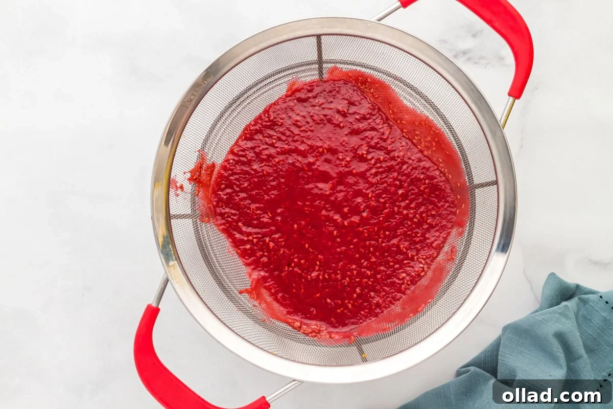 Straining raspberry puree through a fine mesh sieve, separating the smooth sauce from the seeds over a white countertop.