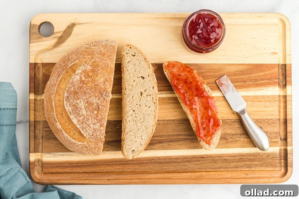 A beautifully baked gluten-free sourdough loaf, perfectly sliced on a rustic wood cutting board, accompanied by a jar of vibrant jam and a knife, showcasing its soft interior and golden crust.