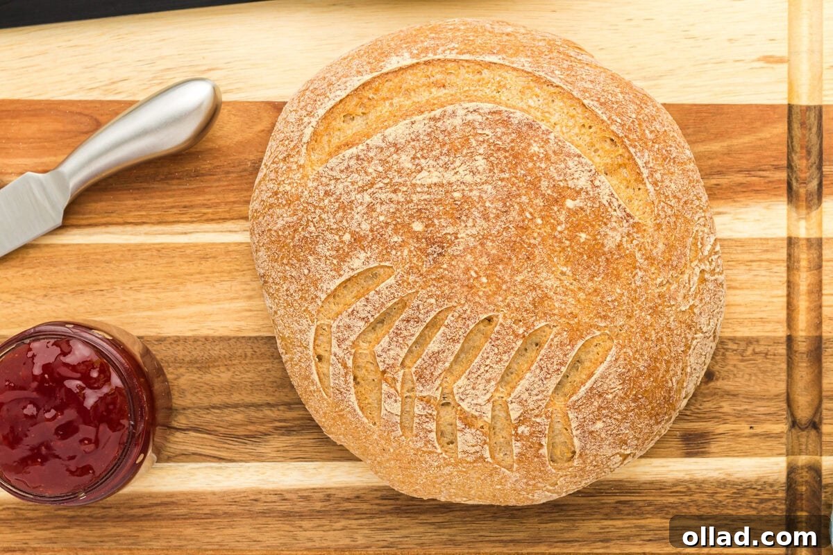 A close-up of a perfectly baked boule of gluten-free sourdough bread, golden brown and crusty, on a wooden cutting board with a knife and jam, ready to be sliced.